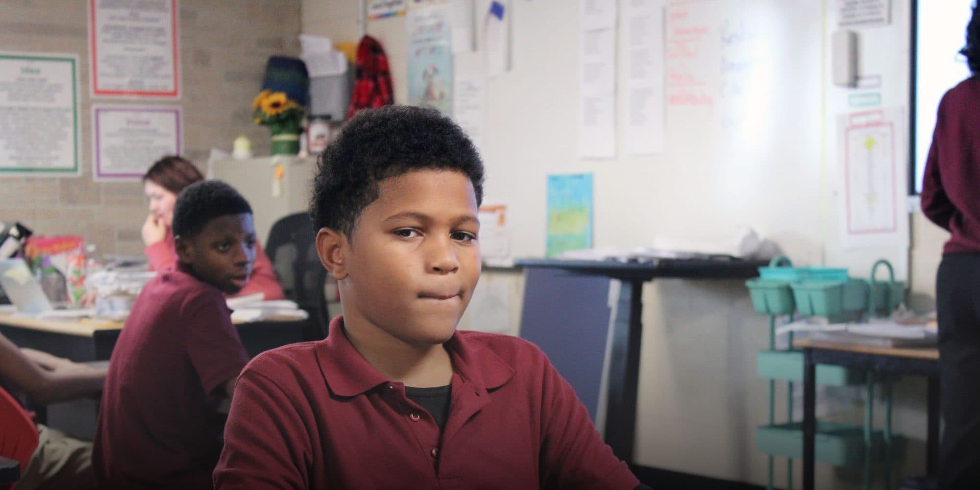 Student in a maroon uniform shirt in a classroom with classmates in the background.