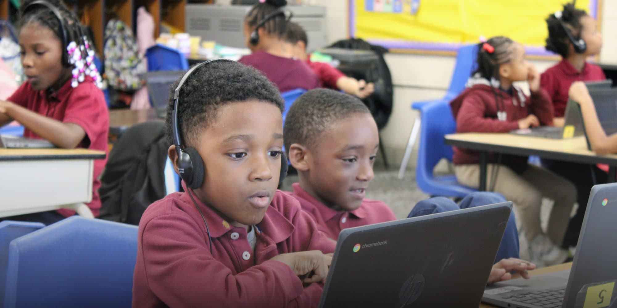 Students in maroon uniforms wearing headphones and working on laptops in a classroom.