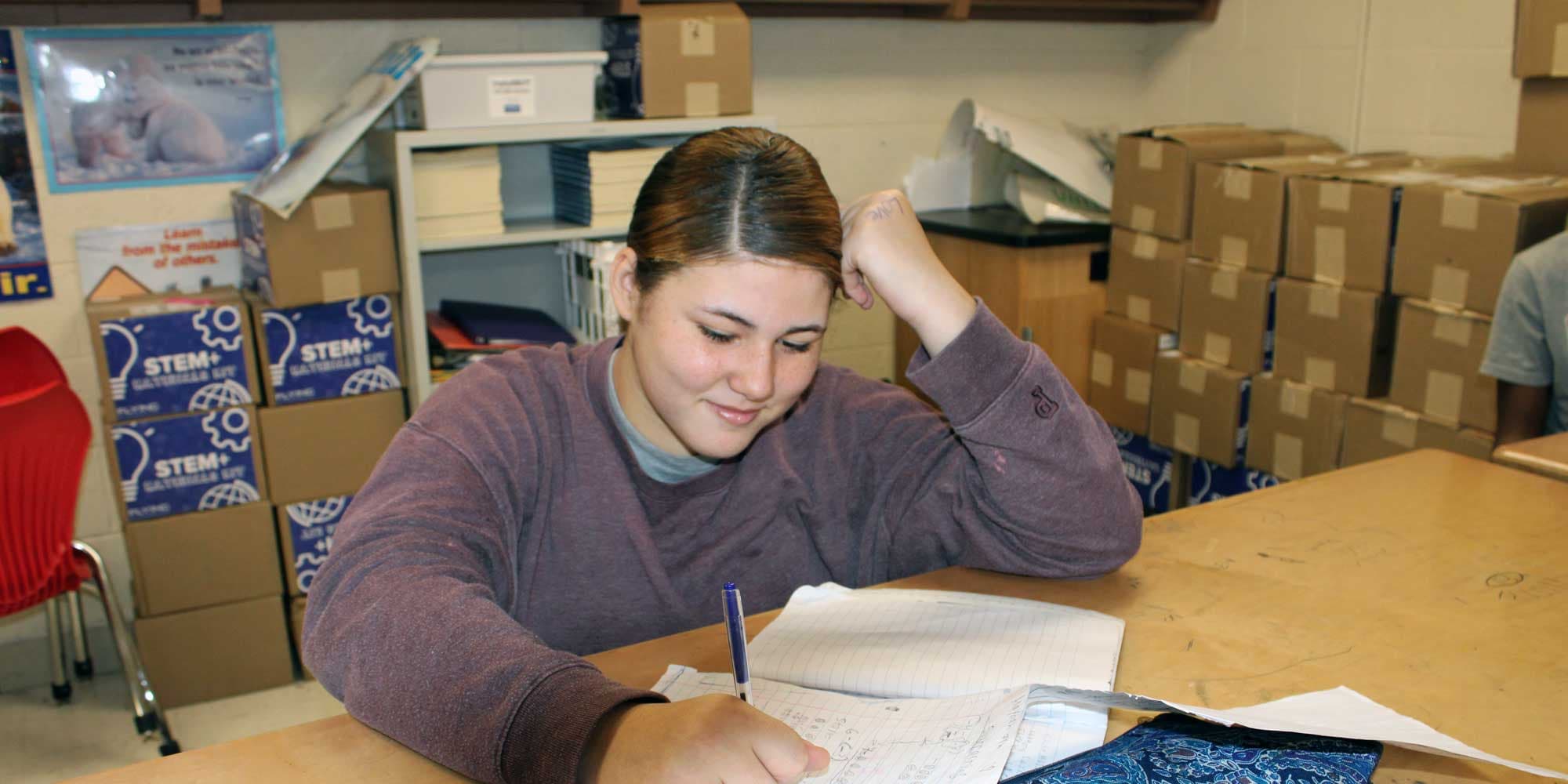 Student writing in a notebook at a classroom table.