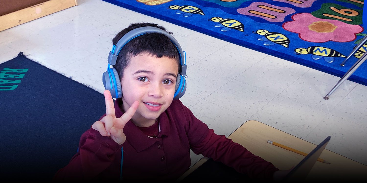 A student sitting in class, wearing a headset and making a peace sign with their hand